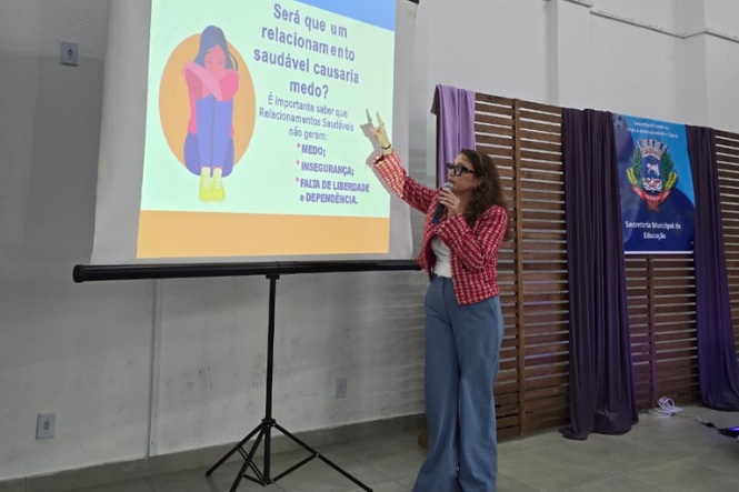 Foto horizontal: mulher fala ao microfone e aponta para apresentação sobre relacionamentos saudáveis, diante de painel da Secretaria Municipal de Educação.
