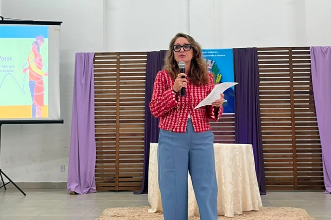 Foto horizontal: mulher fala ao microfone enquanto segura folhas de papel durante palestra sobre relacionamentos saudáveis.
