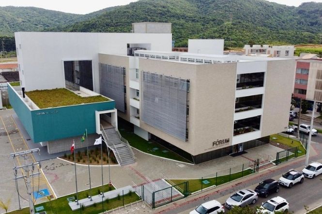 Foto horizontal: vista aérea do Fórum da Comarca de Imbituba, prédio em tons de branco, cinza e azul, com bandeiras e veículos brancos e escuros estacionados ao redor.