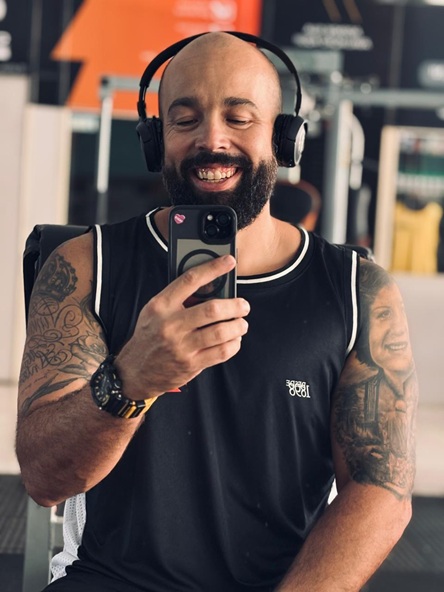Foto vertical: homem sorridente registra selfie no espelho da academia, usando fones de ouvido.