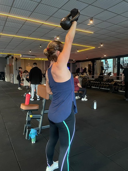 Foto vertical: mulher realiza exercício com kettlebell elevado acima da cabeça em academia.