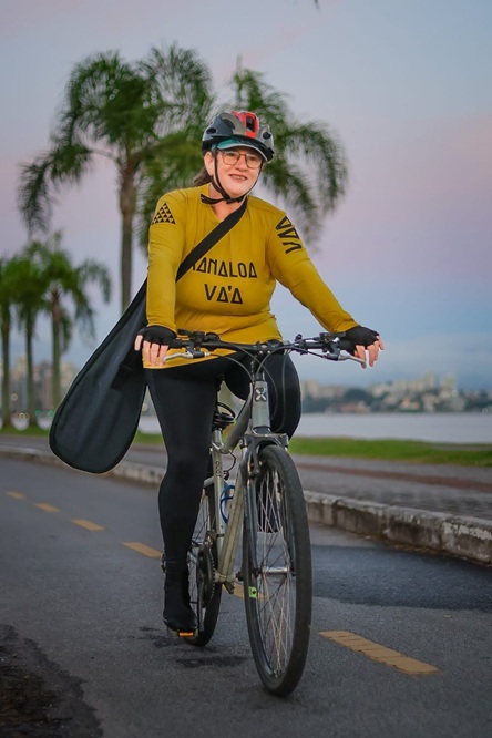 Foto vertical: mulher pedala bicicleta em ciclovia à beira-mar, usando capacete e roupa esportiva.