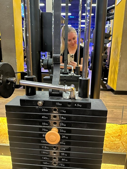Foto vertical: mulher registra selfie pelo espelho diante de aparelho de musculação e pilha de pesos.