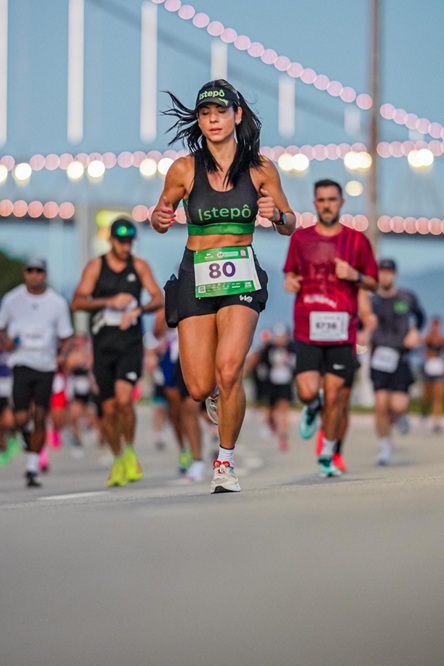 Foto vertical: mulher participa de corrida de rua, com outros competidores ao fundo.