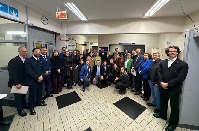 Foto horizontal: grupo de autoridades e servidores posa em ambiente institucional, registro coletivo em corredor interno, clima de integração e cooperação.