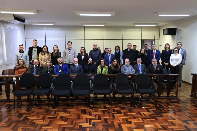 Foto horizontal: grupo de autoridades e servidores posa para registro coletivo em sala de audiência do Fórum.