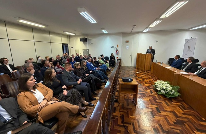 Foto horizontal: público acompanha discurso em auditório durante evento institucional, autoridade fala ao púlpito, plateia atenta, ambiente formal, clima de solenidade e participação.