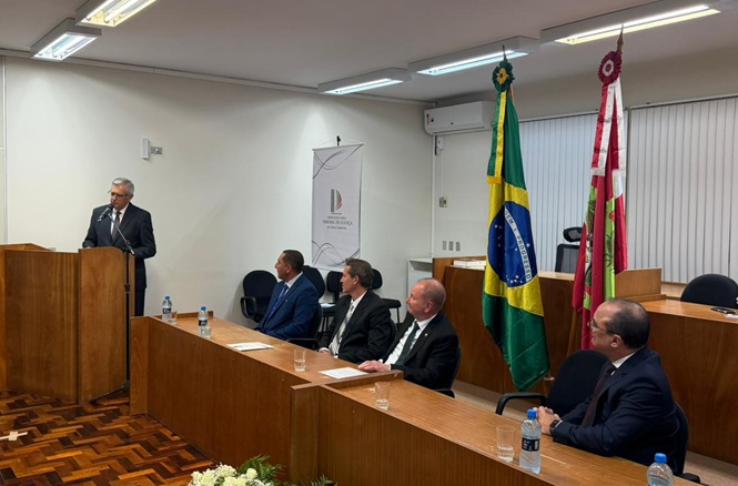 Foto horizontal: autoridade discursa em púlpito durante evento institucional, mesa de autoridades ao lado e bandeiras do Brasil e de SC ao fundo, ambiente formal, clima de solenidade.