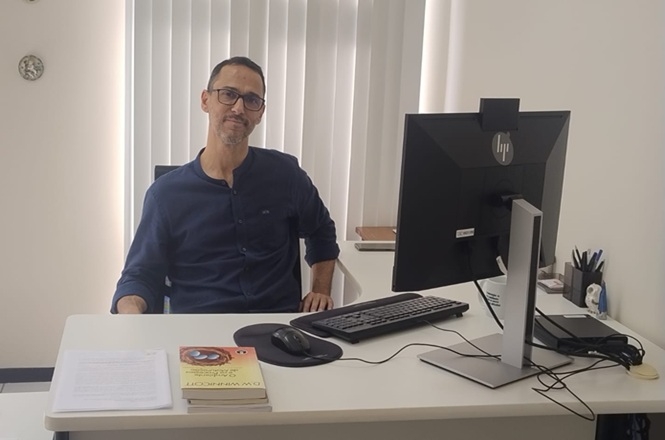 Foto horizontal: psicólogo Leandro Pinheiro de Mello posa para retrato em seu local de trabalho, sentado à mesa diante do computador.