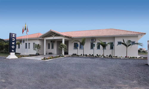 Foto horizontal: fachada do Fórum da comarca de Itapoá, com entrada principal, jardim e mastros com bandeiras.