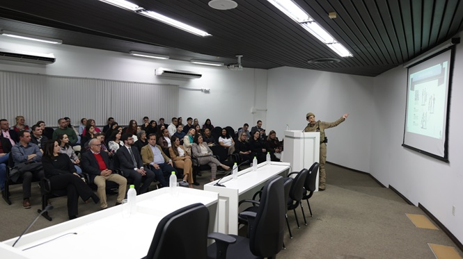 Foto horizontal: policial militar aponta para conteúdo projetado na tela durante palestra para público reunido em auditório.