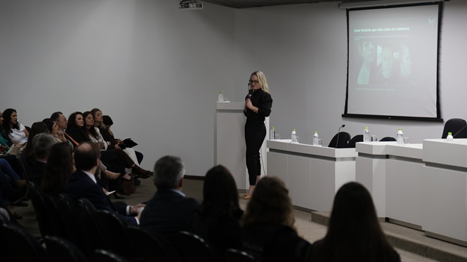Foto horizontal: mulher fala ao microfone diante de público reunido em auditório, com conteúdo projetado em tela ao fundo.