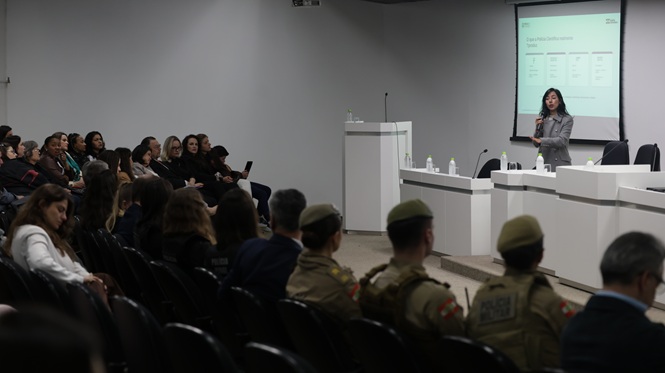 Foto horizontal: mulher apresenta conteúdo diante de público reunido em auditório, com policiais militares entre os participantes.