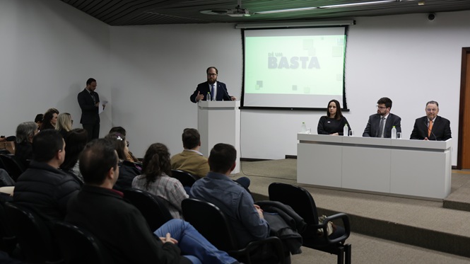 Foto horizontal: homem discursa ao púlpito diante do público, enquanto três integrantes compõem a mesa de autoridades ao lado.