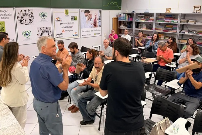 Foto horizontal: grupo participa de atividade em sala de aula, com pessoas em pé conduzindo dinâmica e participantes aplaudindo.
