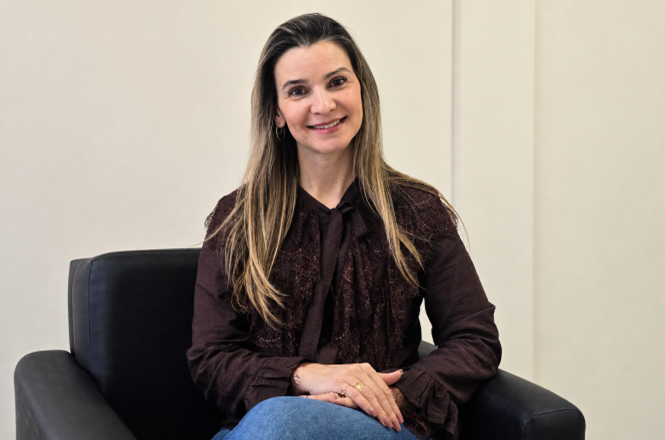 Foto horizontal: psicóloga Joana Patricia Anacleto de Assis posa sorridente para retrato, sentada em uma poltrona e olhando em direção à câmera.
