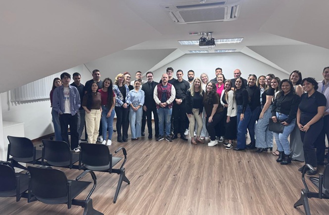Foto horizontal: grupo de participantes posa para foto durante visita institucional, reunido em sala de atividades.