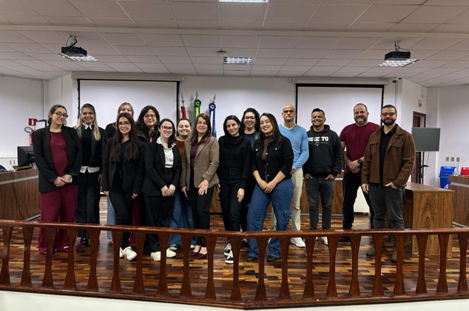 Foto horizontal: grupo de participantes posa para foto em sala de audiência durante visita institucional ao Poder Judiciário.