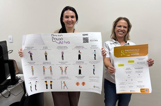 Foto horizontal: duas servidoras sorridentes em trajes branco, preto e calça jeans exibem cartazes informativos nas cores branca, amarela e preta em sala de escritório com paredes claras durante ação de saúde institucional.
