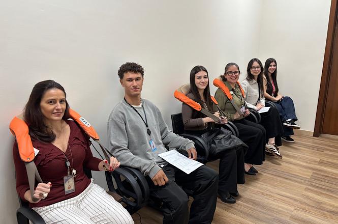 Foto horizontal: grupo de servidores em roupas casuais e formais de tons bordô, cinza, verde e preto senta-se alinhado em cadeiras pretas com aparelhos massageadores laranjas nos ombros em sala de paredes claras e piso amadeirado durante atividade institucional de bem-estar.