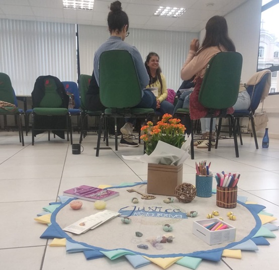 Foto horizontal: participantes sentados em círculo durante encontro de Justiça Restaurativa; em primeiro plano, materiais utilizados na atividade favorecem a escuta, o diálogo e a construção de soluções coletivas.