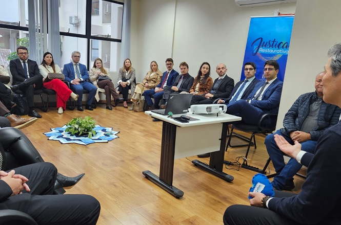 Foto horizontal: reunião institucional sobre justiça restaurativa, participantes sentados em círculo em ambiente interno, banner ao fundo, clima de diálogo e escuta.
