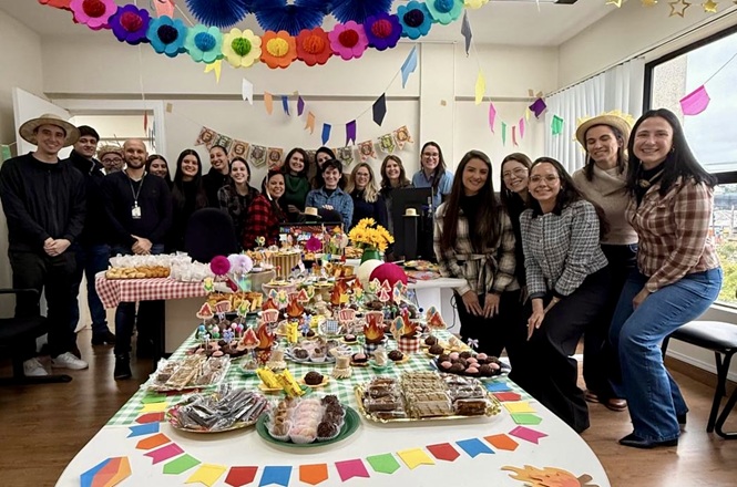 Foto horizontal: Servidores posam ao redor de mesa decorada durante confraternização julina em ambiente de trabalho.