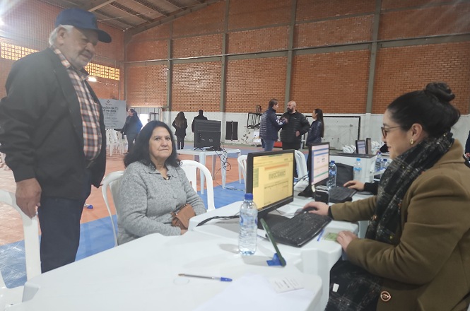 Foto horizontal: atendimento do projeto Justiça até Você em espaço comunitário, equipe utiliza computadores para orientar casal, ambiente acolhedor, tons neutros.