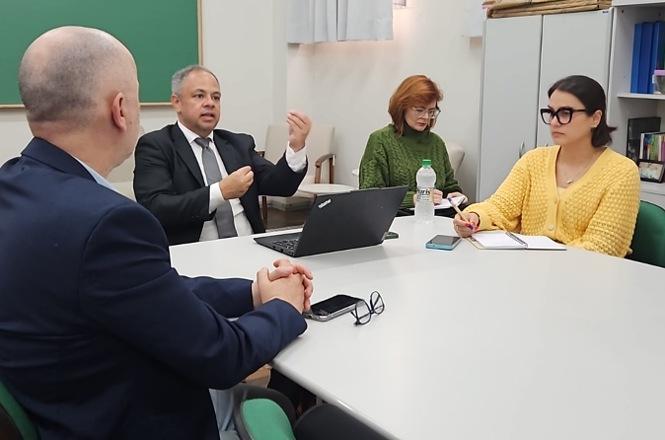 Foto colorida: participantes em reunião à mesa, sala de aula interna, encontro institucional, debatendo propostas em verde, branco e amarelo.