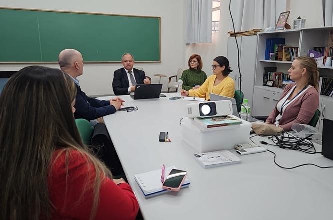 Foto colorida: participantes reunidos à mesa, sala de aula interna, encontro institucional, debatendo propostas em verde, branco e cinza.