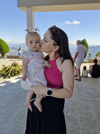 Foto vertical: mulher segura criança no colo e lhe dá um beijo no rosto em ambiente externo.