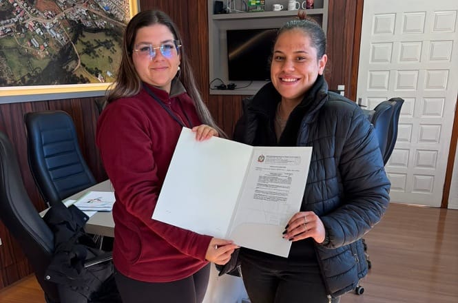 Foto horizontal: servidora e moradora exibem documento de regularização fundiária durante entrega realizada pelo Programa Lar Legal, em ambiente institucional.