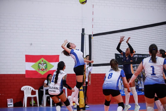 Jogadora salta para ataque em partida de vôlei, bola no alto e equipe em ação, clima de competição e trabalho em equipe