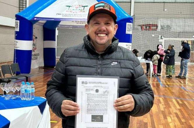 Foto horizontal: homem sorri ao exibir documento durante ação comunitária, ambiente esportivo ao fundo, clima de conquista e cidadania.