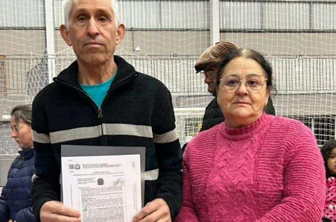 Foto horizontal: casal segura documento em ação comunitária, ambiente esportivo ao fundo, clima de acesso à cidadania.