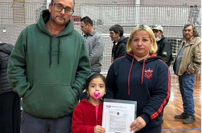 Foto horizontal: família com criança segura documento em ação comunitária, ambiente esportivo ao fundo, clima de cidadania e inclusão.