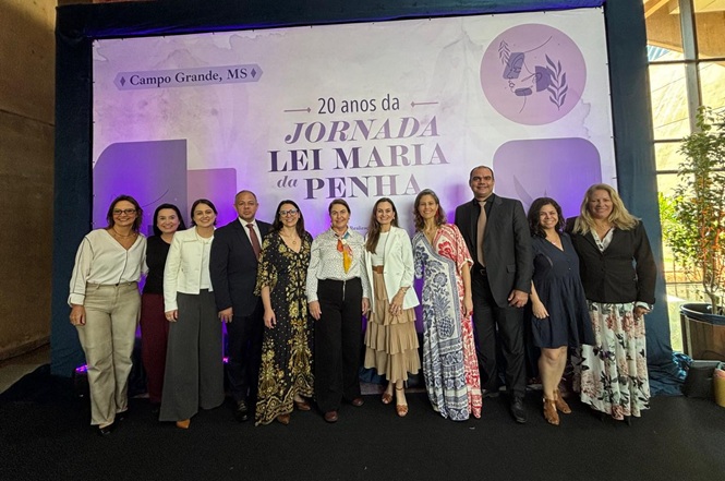 Foto horizontal: grupo de homens e mulheres posa para registro coletivo durante evento alusivo aos 20 anos da Jornada Lei Maria da Penha, em Campo Grande (MS).