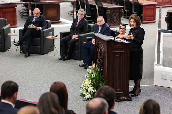 Foto horizontal: autoridade discursa ao público no plenário, enquanto integrantes da mesa acompanham a solenidade.