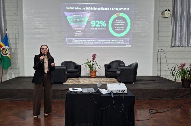 Imagem horizontal mostra palestrante apresentando resultados de 2026 em auditório, com dados projetados ao fundo.