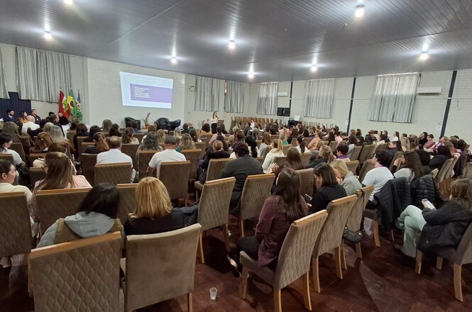 Imagem horizontal mostra público reunido em auditório, acompanhando apresentação projetada em telão.