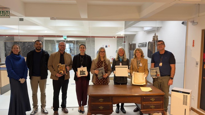 Foto horizontal: grupo posa ao lado de mesa com documentos e peças históricas durante visita ao acervo do Museu do Judiciário Catarinense.
