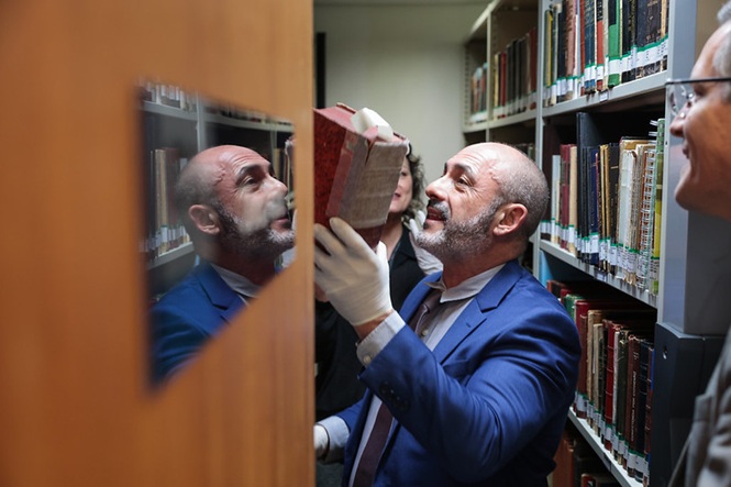 Foto horizontal: 1º vice-presidente do TJSC, desembargador André Luiz Dacol, de terno azul e luvas brancas, manuseia uma obra rara entre estantes de livros da biblioteca.