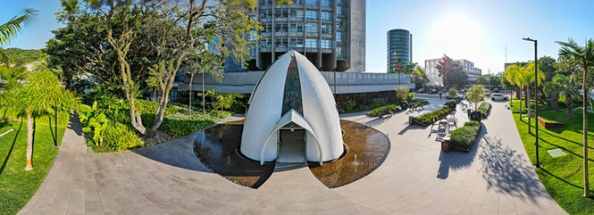 Foto horizontal: capela moderna em área externa do TJSC, jardins e edifícios ao redor, luz natural, tons verdes e claros.