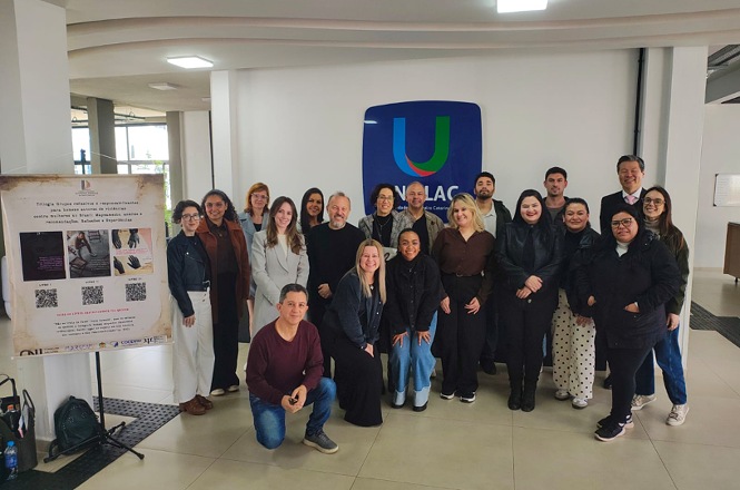 Foto horizontal: grupo de participantes posa para foto em frente à identificação da UNILAC, ao lado de painel informativo.
