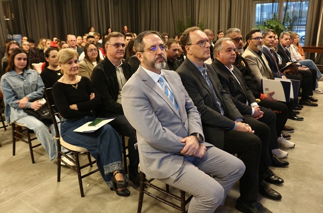 Foto horizontal: autoridades e convidados acompanham cerimônia do programa Novos Caminhos no Alto Vale, sentados em auditório.