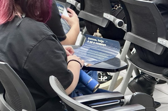 Foto horizontal: jovem sentada em auditório segura material do Novos Caminhos com a frase “Jovem, hoje é só o começo”.