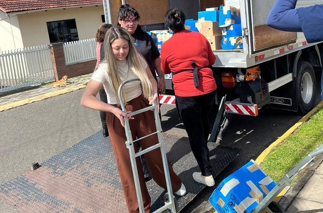 Foto horizontal: mulher movimenta carrinho ao lado de caminhão carregado com caixas azuis e marrons durante transporte.