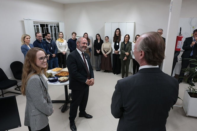 Foto horizontal: de costas, o desembargador Rubens Schulz conversa com o desembargador André Dacol em encontro institucional, grupo acompanha ao fundo, clima de diálogo e integração.