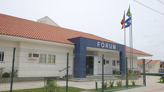 Foto horizontal: fachada do Fórum da comarca de Otacílio Costa, com entrada principal, jardim frontal e mastros com bandeiras.
