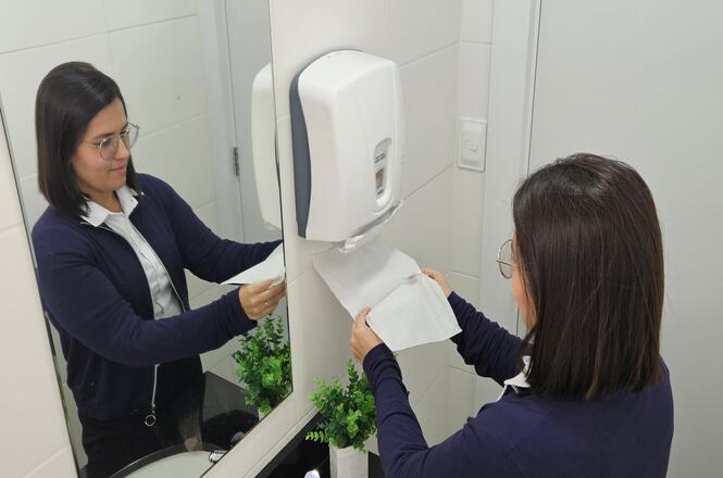 Horizontal: Pessoa retira papel-toalha de dispenser instalado em banheiro institucional, em ambiente com paredes revestidas, espelho amplo, bancada com pia e vaso decorativo ao lado.